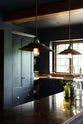 A dramatic view of a Pantry Blue shaker kitchen’s sink run and vintage island topped with Iroko wood. Light stone flooring enhances the space, featuring wooden ceiling accents, a large cooker splashback, and two hanging lights above the island.