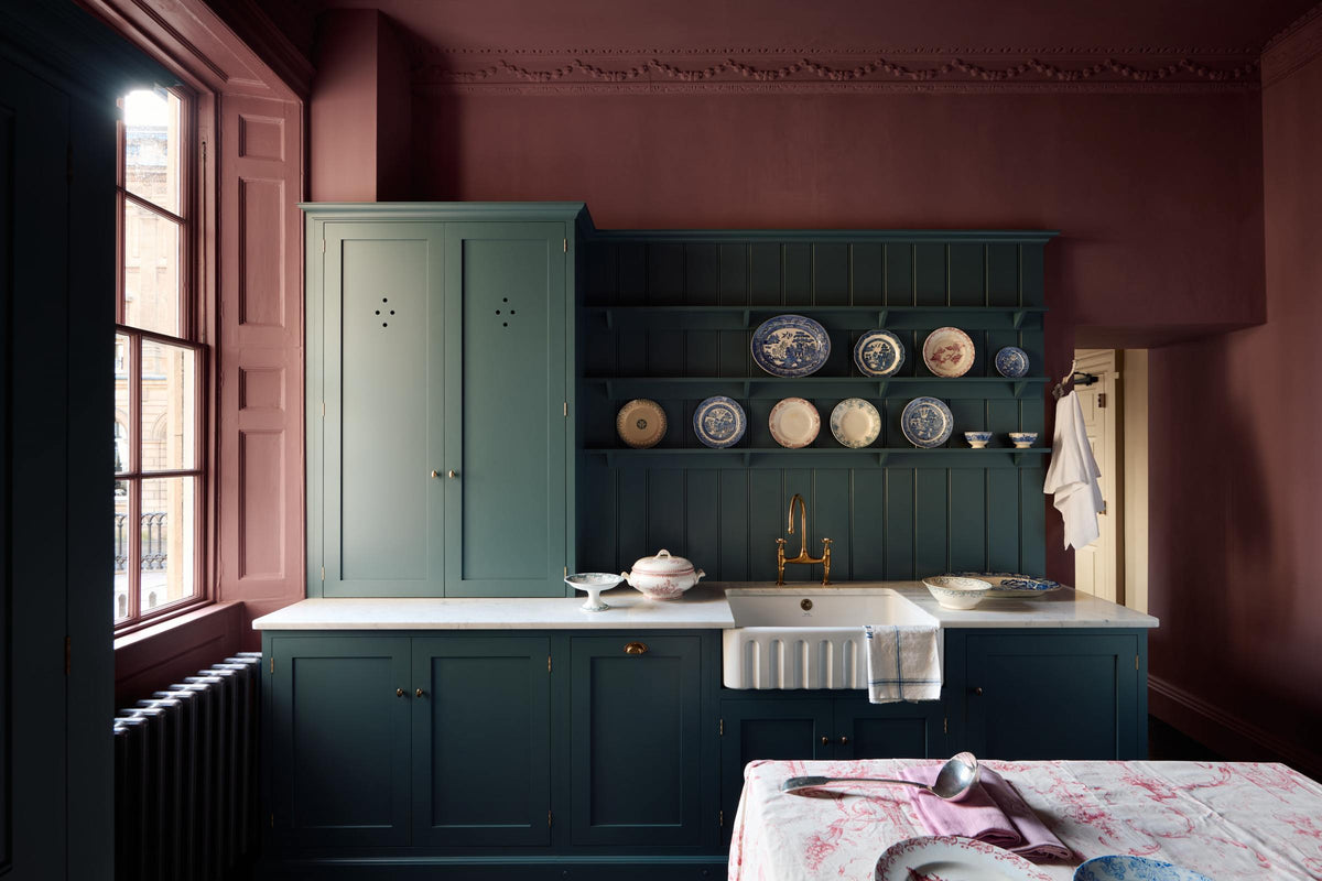 Light streams through sash windows into a moody room with original Georgian features, dusky wine-coloured walls, and dark teal Shaker cabinetry. The display includes a sink run with a fluted ceramic sink, aged brass taps, classic brass door hardware, a white Carrara marble worktop, a tall countertop cupboard, tongue-and-groove wall panelling, and shelves showcasing a selection of classic decorative plates.
