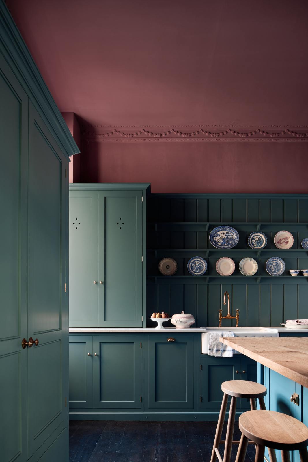 A shaker kitchen’s sink run with a countertop cupboard and panelled wall beside an original sash window. A small island with a rustic oak worktop stands at the centre, accompanied by a row of understated oak Helen’s Stools. Behind them, a grand freestanding pantry from our Classic English range is accented with Victorian-style brass Butler’s Knobs.