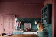A cooker run that is small and compact with Shaker pan drawers on either side of a white Aga range cooker and a large old copper saucepan on the stove. The wall above is adorned with our glossy vintage teal lace market Tiles, an aged brass hanging rail, and a grand London Glazed Wall Cupboard on the adjacent wall. To the left of the run is a door with beautiful original Georgian moulding and architrave, painted in the same dusky wine colour as the walls.