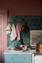 A large display of glossy Lace Market splashback tiles in rich vintage teal contrasts beautifully with wine-coloured walls and aged brass hanging rails adorned with vintage tea towels. In the foreground, a bottle of wine, freshly baked bread, and pastries rest on the Carrara marble worktop of the Clerkenwell Blue Shaker kitchen’s cooker run.