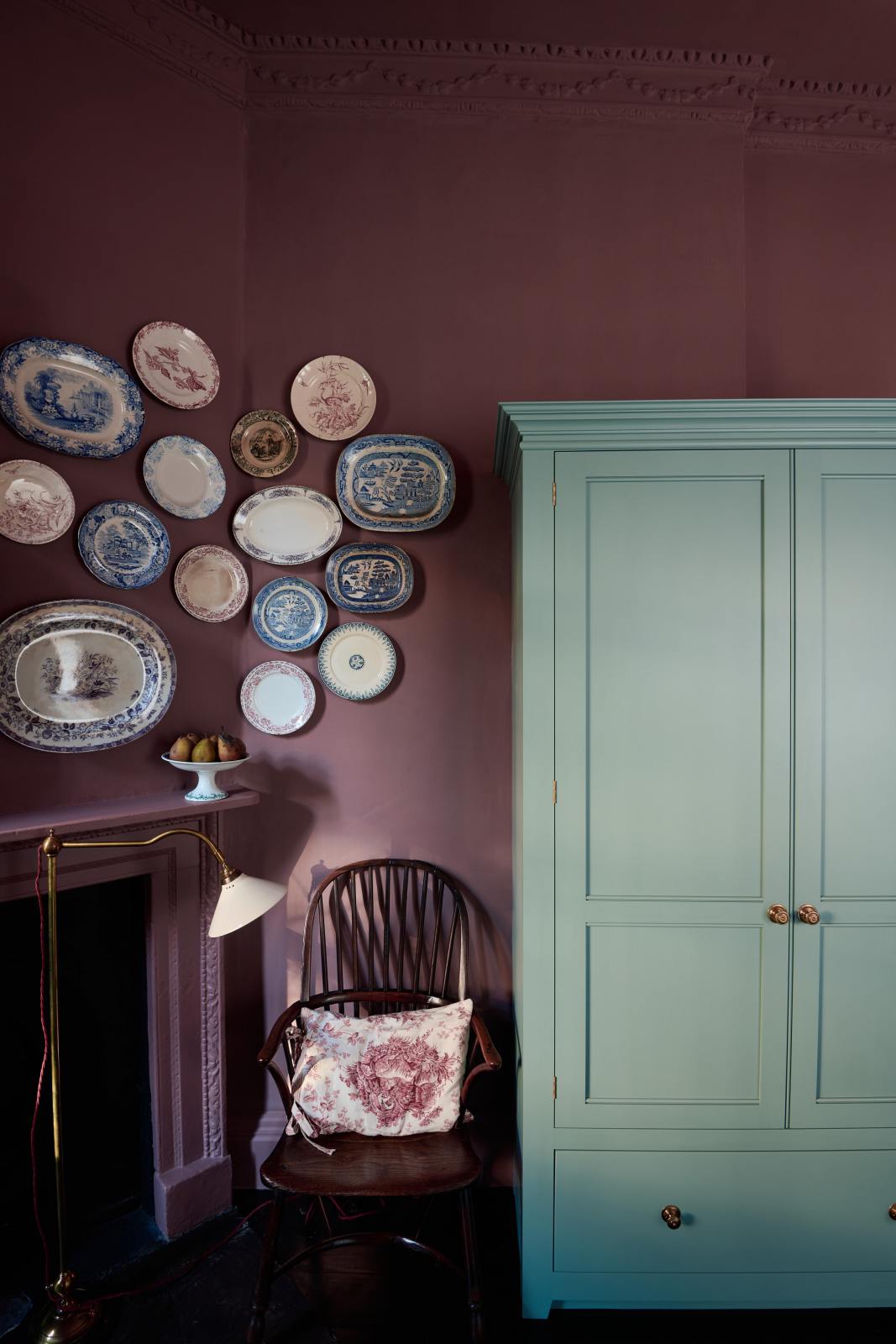 In the far corner of this wine-coloured room, an original Georgian fireplace is paired with a dark, glossy Windsor chair, deVOL’s Grand Tour Reading Lamp, and a collection of vintage Willow pattern plates displayed above the mantlepiece. To the right, a grand freestanding Classic English pantry painted in Clerkenwell blue completes the scene.