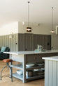 An angled view of a grand warm-grey Shaker kitchen features a cooker run with a Carrara marble worktop, and a wide kitchen island. Adorned with beloved furniture, simple white platters, glass pendant lights, and pale wood herringbone floors, the space feels sophisticated and cool.