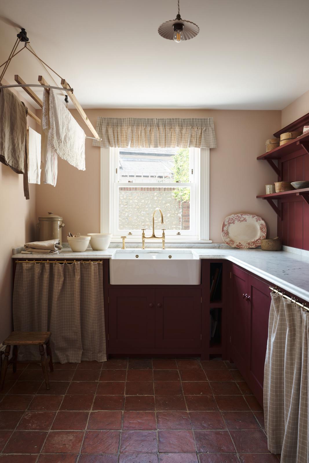 A humble refectory red laundry room features an L-shaped sink run with a Carrara marble worktop, a double Belfast sink with an aged brass Ionian tap, open cabinets covered by grid-patterned curtains on a rail, a laundry maid, and refreshing light from the window.
