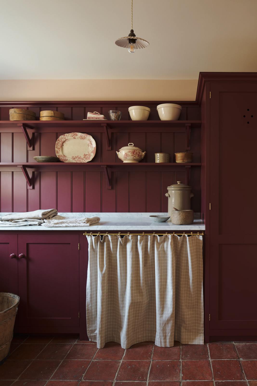 A humble refectory red laundry room features an L-shaped sink run with a Carrara marble worktop, shaker shelves with assorted vintage ceramics, towels, a full-height cupboard, and open cabinets covered by grid-patterned curtains on a rail.