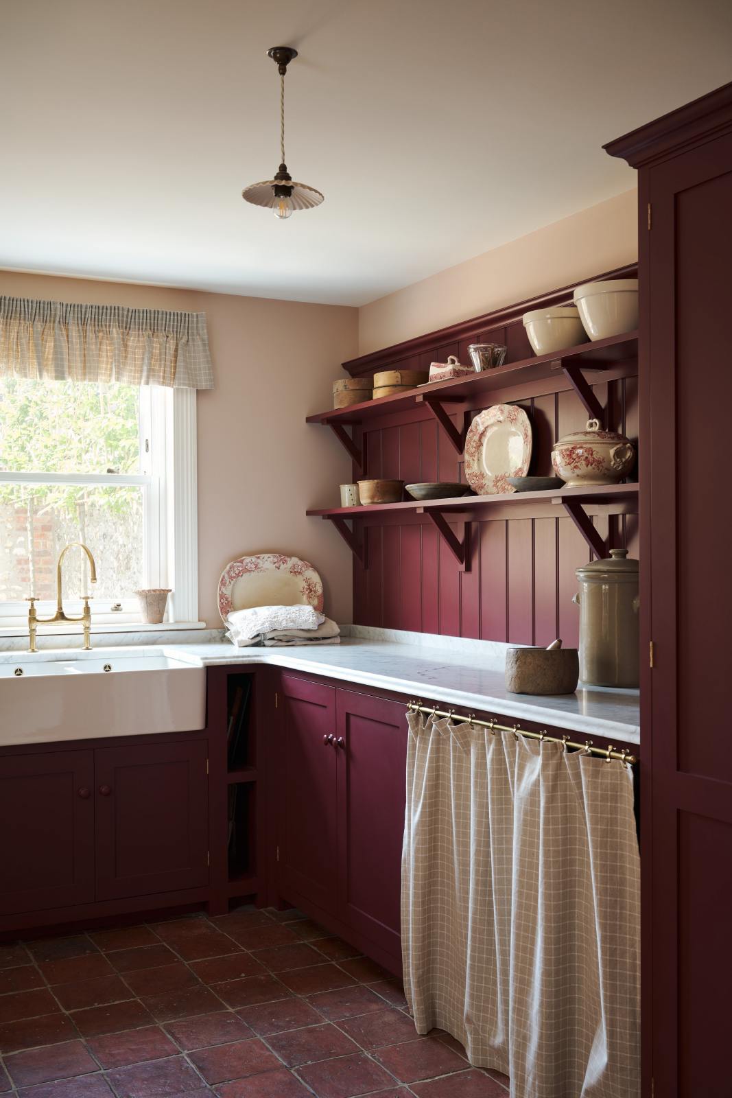 An angled view of a humble refectory red laundry room features an L-shaped sink run with a Carrara marble worktop, a double Belfast sink with an aged brass Ionian tap, open cabinets covered by grid-patterned curtains on a rail, shaker shelves with assorted vintage ceramics, and refreshing light from the window.