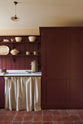 A humble refectory red laundry room features an L-shaped sink run with a Carrara marble worktop, shaker shelves with assorted vintage ceramics, towels, a large full-height cupboard, and open cabinets covered by grid-patterned curtains on a rail.