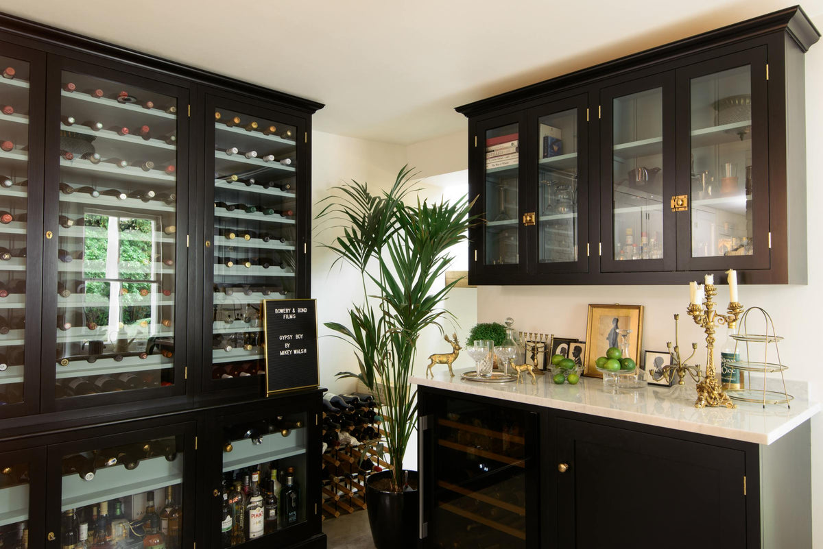 A classy cellar bar features a bank of curiosity cupboards, glazed wall cupboards, and a small wine cooler run with a Carrara marble worktop. Adorned with golden embellishments such as candelabras, a cake stand, deer ornaments, and a golden-framed gentleman's silhouette illustration, it creates a sophisticated and social space.