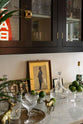 A closer look at a classy cellar bar’s glazed wall cupboards and a small wine cooler run with a Carrara marble worktop. Adorned with golden embellishments such as candelabras, deer ornaments, a golden-framed gentleman's silhouette illustration, and vintage red Tabac neon lights, it creates a sophisticated and social space.