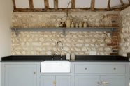 A chic and simple muted-blue sink run against the original wall textures of the pool house features a single Belfast sink with a nickel Ionian tap, a black honed granite worktop, and a long shelf. Adorned with vintage ceramics, brass fittings, and vintage steel baskets, it creates a functional corner in the space.