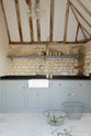 A chic and simple muted-blue sink run against the original wall textures of the pool house features a single Belfast sink with a nickel Ionian tap, a black honed granite worktop, and a long shelf. Adorned with vintage ceramics, brass fittings, and vintage steel baskets, it creates a functional corner in the space.