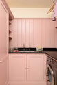 A chic and simple muted-pink laundry room features a sink run with a honed black granite worktop, a full-height cupboard, a single sink, and nickel Mayan taps. Adorned with empty jam jars, folded tea towels, and a black washing
machine, it creates a functional corner in the space.