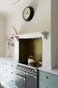 An angled view of the minimal Trinity Blue shaker kitchen features a cooking area with a marble countertop, designed around a chimney breast, and complemented by a nearly golden-finish copper splashback. In the kitchen, you'll find a classic round clock with a black rim, assorted ceramics, glass vases filled with wildflowers, and a cream kettle sitting on the stove.