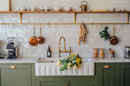 A close-up of the extra-large double fluted Belfast sink with an aged brass tap as part of the beautiful wilkes green painted sink run. The shimmering, glossy beige tiles of the backsplash perfectly complement the aged brass hanging rails and classic shelf brackets.