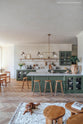 A spacious, open plan room with high ceilings, textured parquet wooden floors and a beautiful wilkes green painted shaker kitchen. The walls are painted in a creamy, limewash paint and the backsplash is fitted with glossy beige tiles. A large kitchen island dominates the image, with four Helen’s Stools pulled up to it. The room feels fresh and contemporary but still has lots of traditional charm.