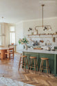 An angled view of a spacious, open plan room with high ceilings, textured parquet wooden floors and a beautiful wilkes green painted Shaker kitchen. The walls are painted in a creamy, limewash paint and the backsplash is fitted with glossy beige tiles. A large kitchen island dominates the image, with four Helen’s Stools pulled up to it. The room feels fresh and contemporary but still has lots of traditional charm.