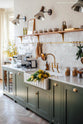 An oblique view of the sink run of a beautiful wilkes green painted shaker kitchen featuring textured parquet wooden floors. The walls are painted in a creamy, limewash paint, and the backsplash is fitted with glossy beige tiles.  The room feels fresh and contemporary but still has lots of traditional charm.