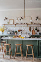 An angled view of a spacious, open plan room with high ceilings, textured parquet wooden floors and a beautiful wilkes green painted shaker kitchen. The walls are painted in a creamy, lime-wash paint and the backsplash is fitted with glossy beige tiles. A large kitchen island dominates the image, with four Helen’s Stools pulled up to it. The room feels fresh and contemporary but still has lots of traditional charm.