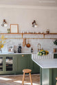 A close-up, angled view of a spacious, open plan room with high ceilings, textured parquet wooden floors and a beautiful wilkes green painted shaker kitchen. The walls are painted in a creamy, limewash paint and the backsplash is fitted with glossy beige tiles.