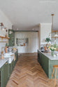 A spacious view of a beautiful wilkes green painted shaker kitchen with textured parquet wooden floors overlooking a mushroom off-white pantry and storage with shelves and a wooden step ladder.