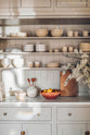 A closer look at the bespoke mushroom off-white pantry reveals shelves and a marble countertop. It features a collection of beautifully minimalistic glazed ceramics, books, wooden utensils, a wooden chopping board, and a maroon bowl of lemons.
