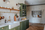 The spacious open plan room features high ceilings, textured parquet wooden floors, and a charming ‘Wilkes Green’ painted Shaker kitchen. The creamy limewash walls complement the glossy beige tile backsplash. A bespoke off-white pantry with shelves and a marble countertop holds minimalistic glazed ceramics, books, and a wooden chopping board, creating a fresh yet traditionally charming atmosphere.