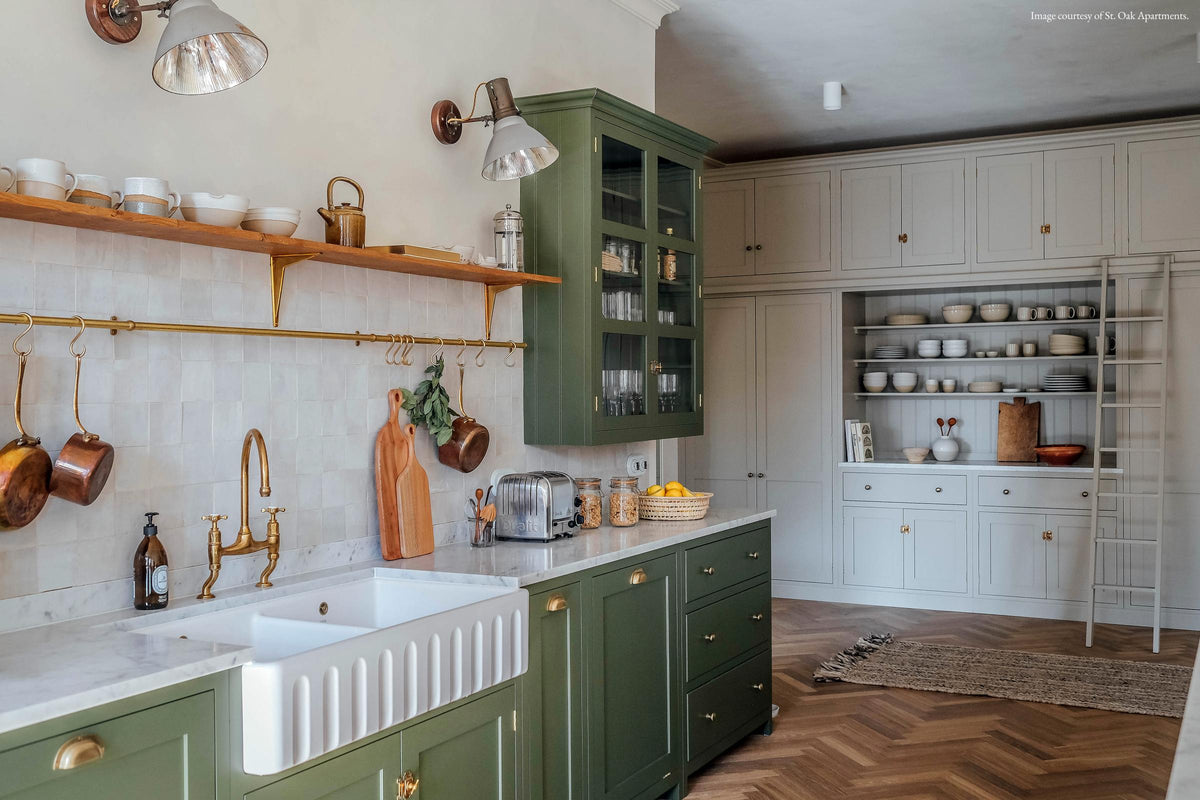 The spacious open plan room features high ceilings, textured parquet wooden floors, and a charming ‘Wilkes Green’ painted Shaker kitchen. The creamy limewash walls complement the glossy beige tile backsplash. A bespoke off-white pantry with shelves and a marble countertop holds minimalistic glazed ceramics, books, and a wooden chopping board, creating a fresh yet traditionally charming atmosphere.