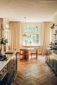 A warm, glowy image of a mid-century style dining area at the bottom of a traditional Shaker kitchen. A large window at the back of the room creates a pool of beautiful, summery light whilst wooden parquet floors, limewash walls and floor-to-ceiling curtains add layers of texture.