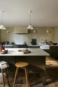 An olive green Shaker kitchen featuring a sink and cooker run, a freestanding prep table with a Carrara marble worktop, and Helen Stools gathered around the breakfast nook. A large wooden shelf displays a thoughtful selection of muted ceramics and mugs. The space embraces a simple theme rooted in raw, natural materials—an ode to Mother Nature—highlighted by a small oil painting of trees placed beside the single Belfast sink with an aged brass Ionian tap.