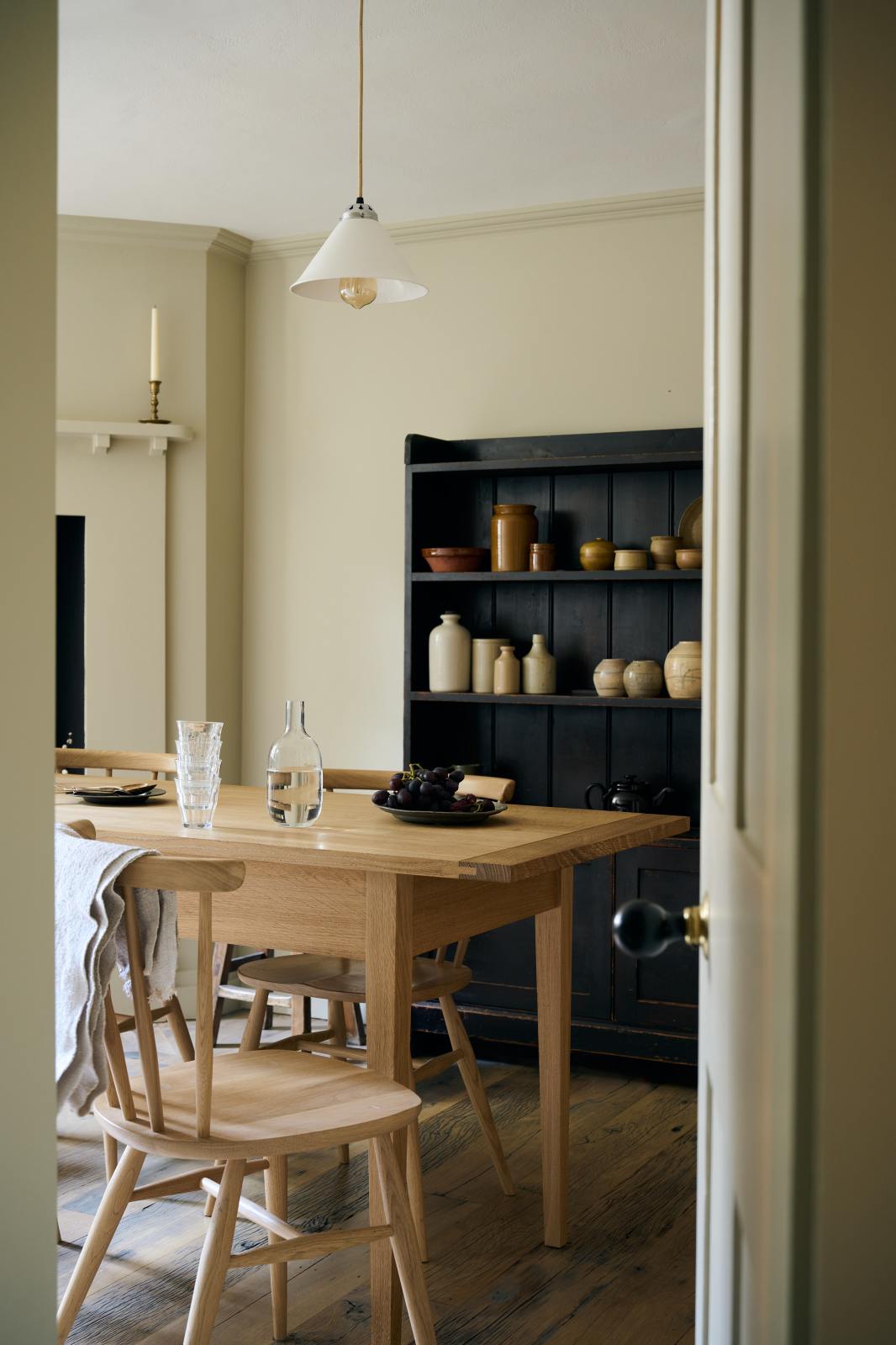 Looking through the open white door, a pale wooden dining table with matching chairs comes into view. Behind it, a freestanding open larder displays beautifully simple cream, green, and reddish-brown ceramic vases and jugs.