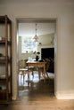 Looking through the archway in the kitchen, beside a large wooden shelf displaying a thoughtful selection of muted ceramics and mugs, the dining room comes into view with a pale wooden dining table and matching chairs. A large open fireplace adds warmth to the space, accompanied by a generous storage cupboard and cabinets. A freestanding open larder showcases beautifully simple cream, green, and reddish-brown ceramic vases and jugs.