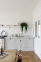 A beautiful Trinity Blue Shaker kitchen featuring a cooker and sink run, a vintage wooden dining table with a wonderfully worn top, and a single copper sink with aged brass Mayan taps. A long Shaker shelf spans above the kitchen area, complemented by a low, hanging house plant, antique furniture, and grand solid oak herringbone floors.