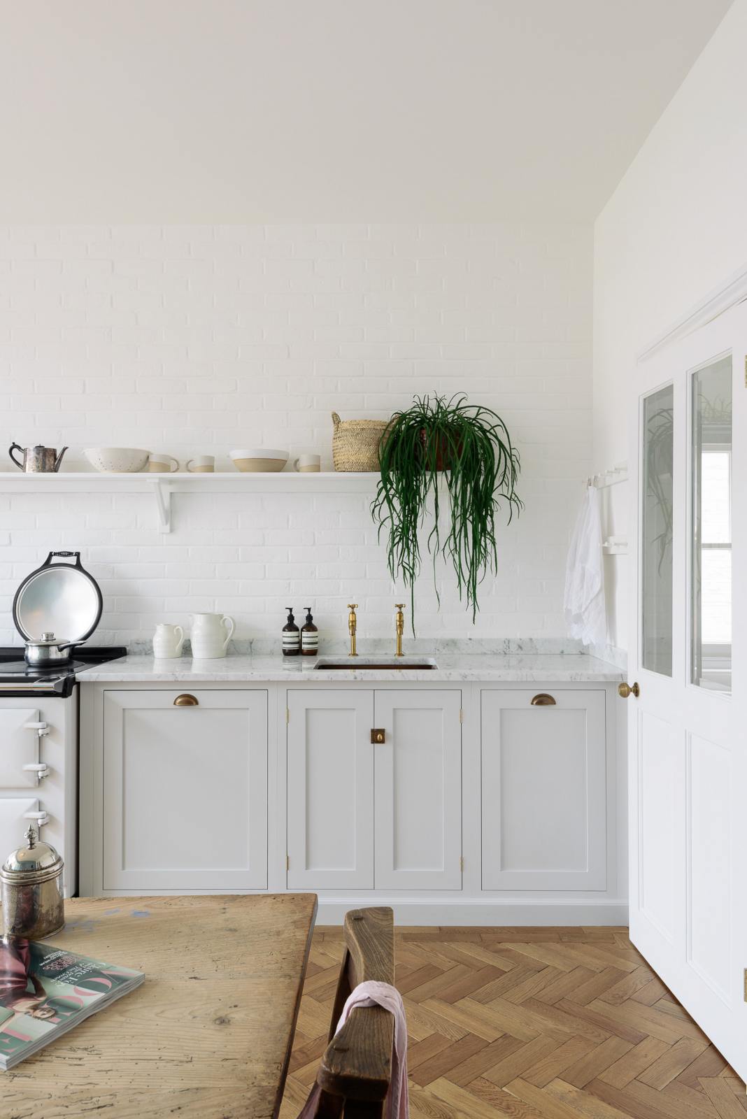 A beautiful Trinity Blue Shaker kitchen featuring a cooker and sink run, a vintage wooden dining table with a wonderfully worn top, and a single copper sink with aged brass Mayan taps. A long Shaker shelf spans above the kitchen area, complemented by a low, hanging house plant, antique furniture, and grand solid oak herringbone floors.
