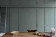A close-up of a beautiful Trinity Blue Shaker kitchen featuring floor-to-ceiling pantry cabinets with a matching ladder, and a vintage wooden dining table with a wonderfully worn top. The table has a collection of baking equipment and ingredients.
