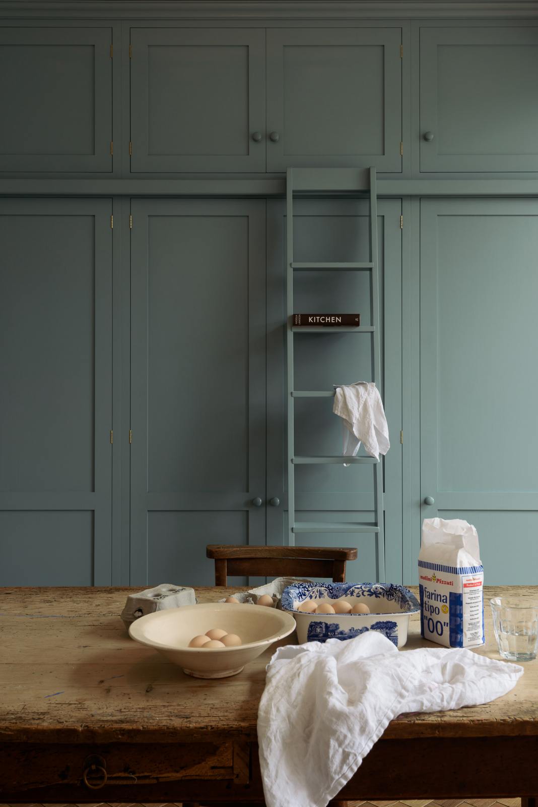 A close-up of a beautiful Trinity Blue Shaker kitchen featuring floor-to-ceiling pantry cabinets with a matching ladder, and a vintage wooden dining table with a wonderfully worn top. The table has a collection of baking equipment and ingredients.