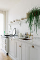 An oblique and close-up view of a beautiful Trinity Blue Shaker kitchen featuring a cooker and sink run, a white gas stove and a single copper sink with aged brass Mayan taps. A long Shaker shelf spans above the kitchen area, complemented by a draped potted plant and grand solid oak herringbone floors.