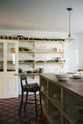 An atmospheric shot of an antique green leather upholstered bar stool, propped up at a large Shaker island in the centre of a utilitarian-looking scullery. Behind the island, four rows of Shaker shelves, painted in a pale yellow hue, are fitted to a wall of glossy white subway tiles, a soft contrast to the original red and black chequerboard quarry tile floor.