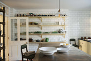 A landscape shot of a moody and atmospheric butter-yellow painted shaker kitchen with a simple, utilitarian feel. Light cupboards and glossy, white tiled walls contrast with original black and red chequerboard quarry tiles. The scullery features a central island, a long run of base cabinets that includes a double sink unit, honed back granite worktops and fitted Shaker shelving.