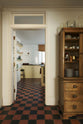 Looking through a doorway into a basement scullery with Shaker base cupboards and a central island, all painted in a soft yellow hue and fitted with a combination of honed black granite and reclaimed wooden worktops. Floor-to-ceiling glossy subway tiles create beautiful reflections, contrasting with the time-worn, original black and red chequered quarry tiles.