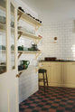 An angled view towards a corner of shaker base cabinets and four fitted shaker shelves, all painted in a soft creamy yellow colour. Glossy white tiled walls and original, black and red quarry tiled floors create a utilitarian feel.