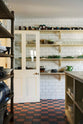 Four rows of Shaker shelves, painted in a muted, buttery yellow hue, are fitted to a white floor-to-ceiling tiled wall, and are stocked with jars of jam, glassware, bowls and vibrant green majolica leaf pottery. This scullery kitchen, found below stairs in a grand Surrey country house, feels very utilitarian with glossy subway tiles and original Victorian red and black chequered quarry tiles.