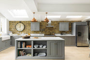 A soft industrial grey Shaker kitchen features an L-shaped sink and cooker run with a Carrara marble worktop, a kitchen island, wall cupboards, and a large silver refrigerator. Adorned with a large pocket watch clock, a weathered brick wall installation, copper pendant lights, stacks of colourful books, and basic kitchen necessities, it creates a modern and functional kitchen for enthusiastic food connoisseurs.