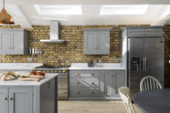 A soft industrial grey Shaker kitchen features an L-shaped sink and cooker run with a Carrara marble worktop, a kitchen island, wall cupboards, and a large silver refrigerator. Adorned with a steel canopy, a weathered brick wall installation, a cream gas cooker, and a stone pestle and mortar, it creates a modern and functional kitchen for enthusiastic food connoisseurs.