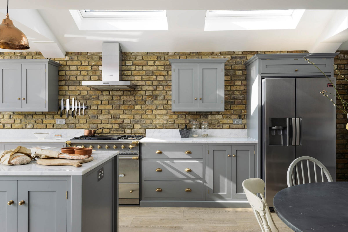 A soft industrial grey Shaker kitchen features an L-shaped sink and cooker run with a Carrara marble worktop, a kitchen island, wall cupboards, and a large silver refrigerator. Adorned with a steel canopy, a weathered brick wall installation, a cream gas cooker, and a stone pestle and mortar, it creates a modern and functional kitchen for enthusiastic food connoisseurs.