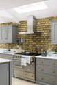 An oblique view of a soft industrial grey Shaker kitchen features an L-shaped sink and cooker run with a Carrara marble worktop, a kitchen island, wall cupboards, and a large silver refrigerator. Adorned with a steel canopy, a weathered brick wall installation, a cream gas cooker, and a stone pestle and mortar, it creates a modern and functional kitchen for enthusiastic food connoisseurs.