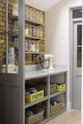 The edge of a soft industrial grey Shaker kitchen features a small walk-in pantry cupboard with open base cabinets and shelves. Adorned with a weathered brick wall installation, dried ingredients in glass jars, and a mixing machine, it creates a modern and functional kitchen for enthusiastic food connoisseurs.