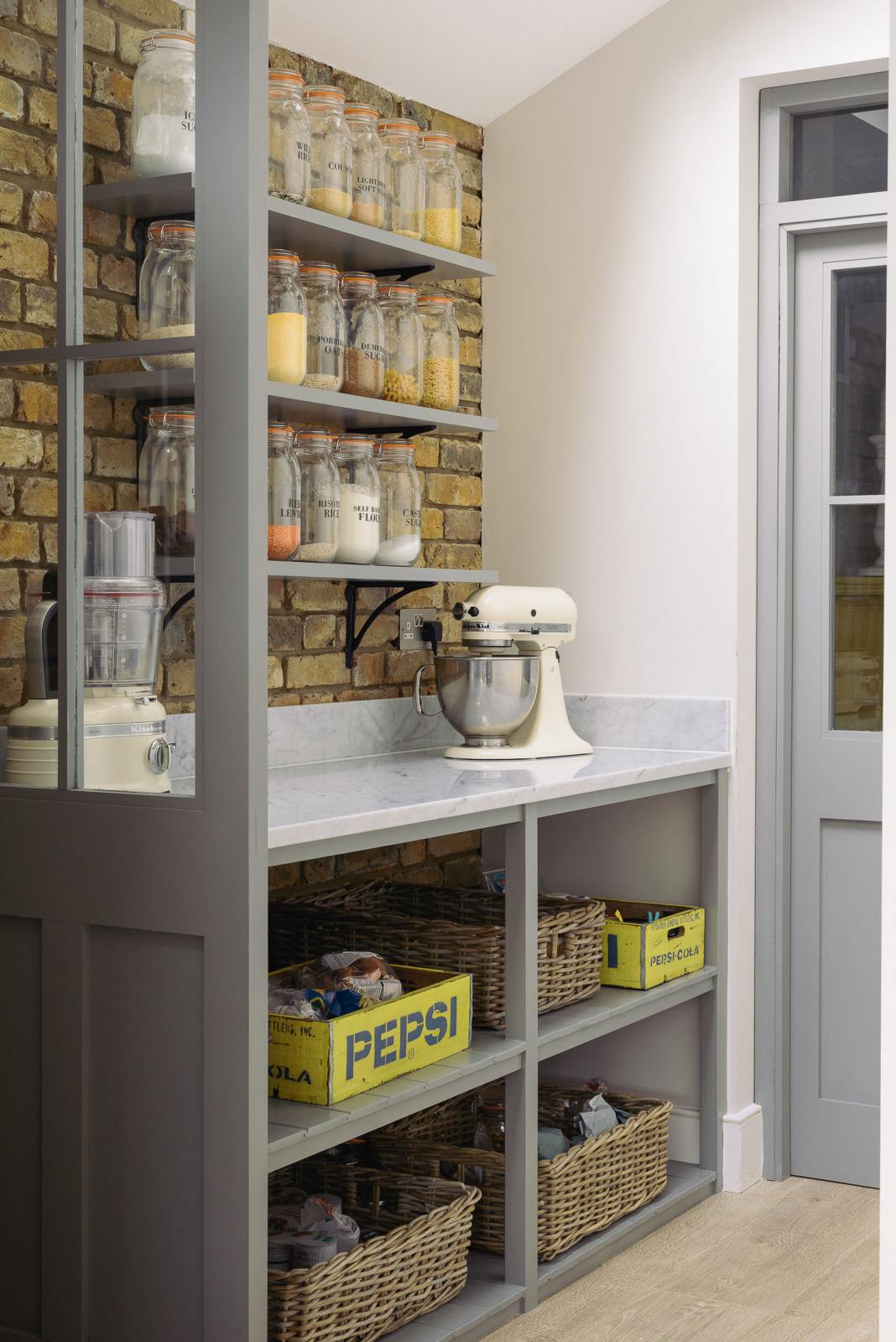 The edge of a soft industrial grey Shaker kitchen features a small walk-in pantry cupboard with open base cabinets and shelves. Adorned with a weathered brick wall installation, dried ingredients in glass jars, and a mixing machine, it creates a modern and functional kitchen for enthusiastic food connoisseurs.