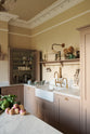 Past the large kitchen island of a romantic and peaceful Princelet pink Shaker kitchen is a tactile corner of a sink run, an open countertop cupboard, shelf with brass railings, textured marble worktop, and a fridge and freezer cupboard. Beautifully paired with the subtle butter yellow painted walls, decorative cornice, beautiful mugs and assorted vintage personal items, it creates a warm and inviting space.