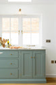 A corner of a bright and elegant Trinity blue Shaker kitchen features a long cooker run with a Carrara marble worktop. Beautifully decorated with artisan ceramics, books, wooden chopping boards, sweet potatoes, onions, and garlic cloves, it creates a humble family space.