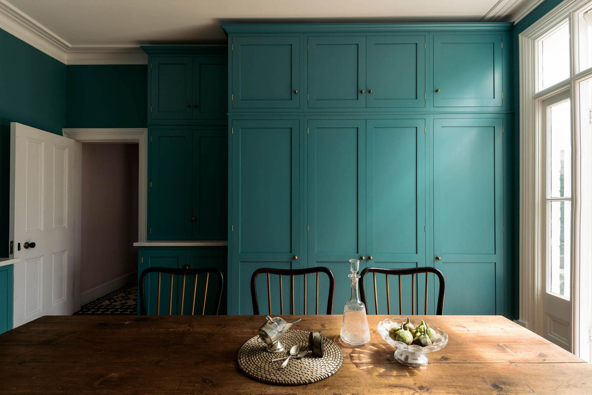 A grand, seafoam green Shaker kitchen featuring a bank of full-height pantry cupboards and a free-standing closed larder with Carrara marble worktop. Adorned with beautiful vintage furniture, a glass bowl of green figs, antique silverware, and a decanter, the kitchen reflects the refined tastes of its socialite and collector owners.